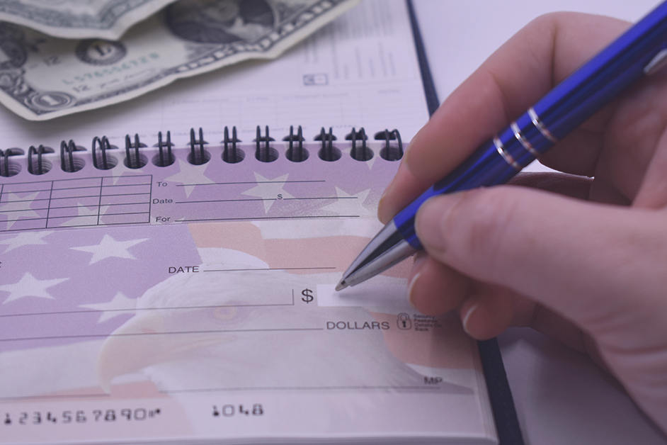 A close-up of a hand holding a blue pen, filling out a check in a spiral-bound checkbook, with U.S. dollar bills visible in the background.